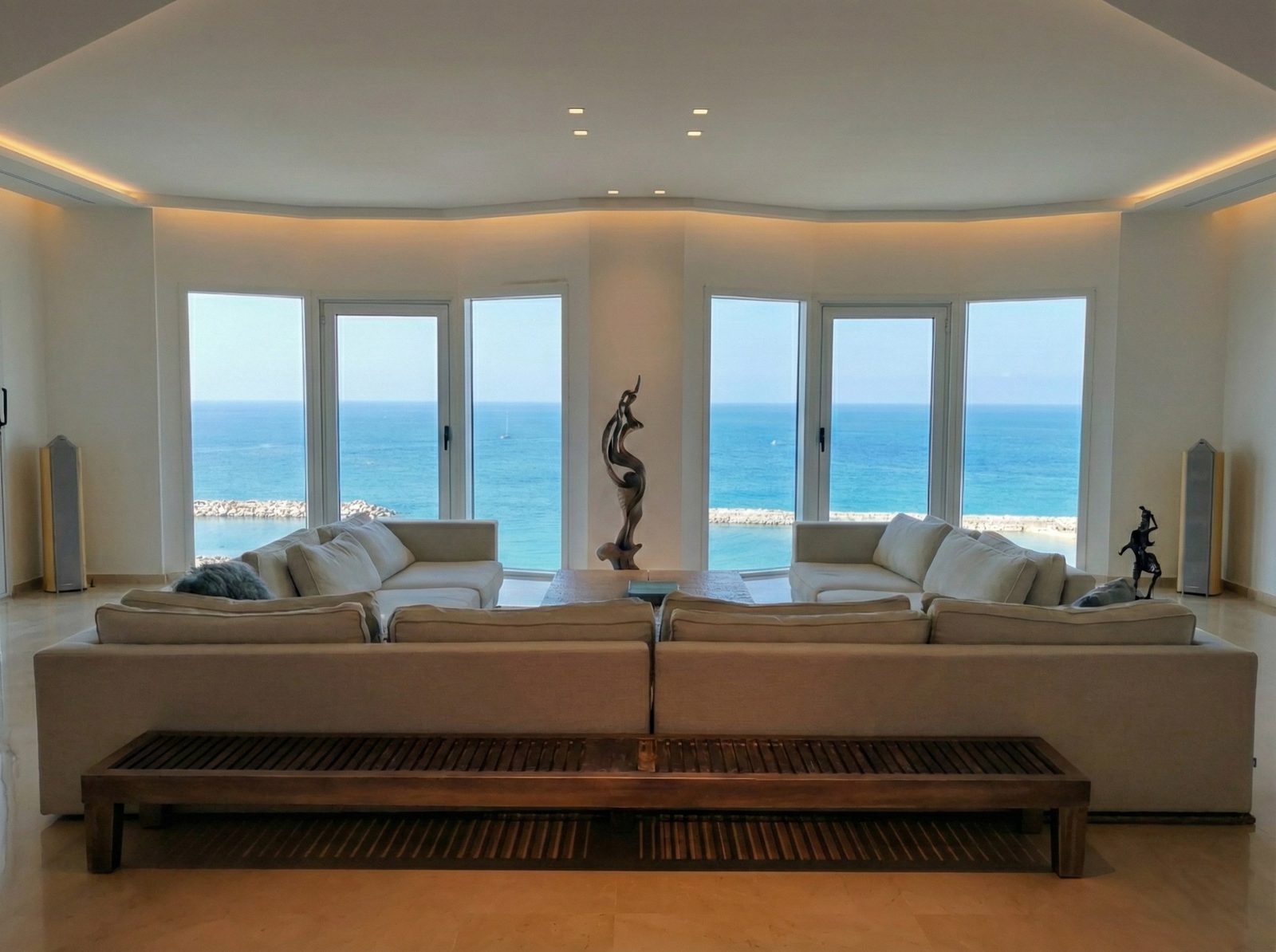 Living room of the Herzliya Pituach apartment: a tall bronze toned abstract sculpture standing between two runs of windows that open onto the sea, cream sofas and a slatted wooden bench in front